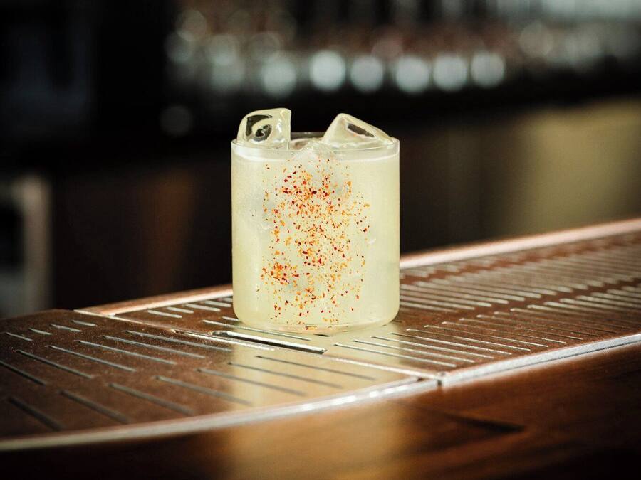 A bar setting with a glass of a beverage garnished with a red spice, a metal bar counter, and a blurred background of bottles.