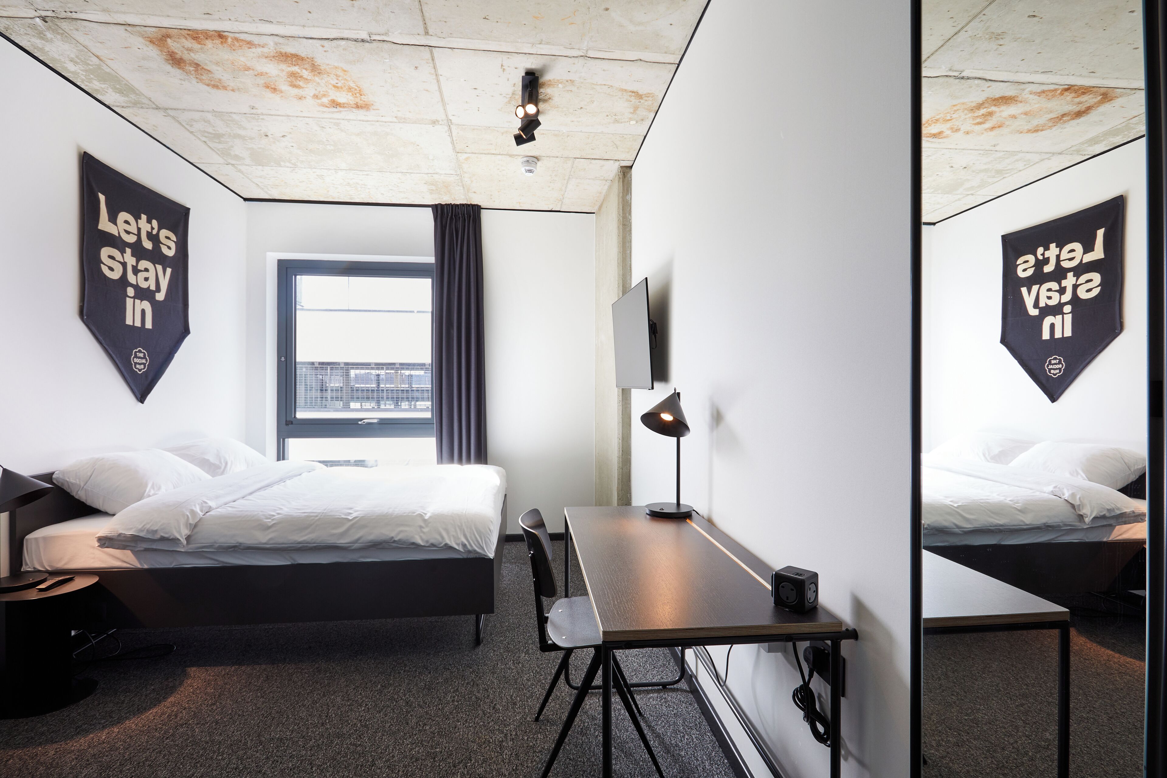 A modern hotel room with a bed, desk, and chair, a window with curtains, and two motivational banners on the walls.