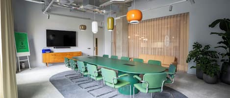 A conference room with a long table, green chairs, a television, a whiteboard, and potted plants.