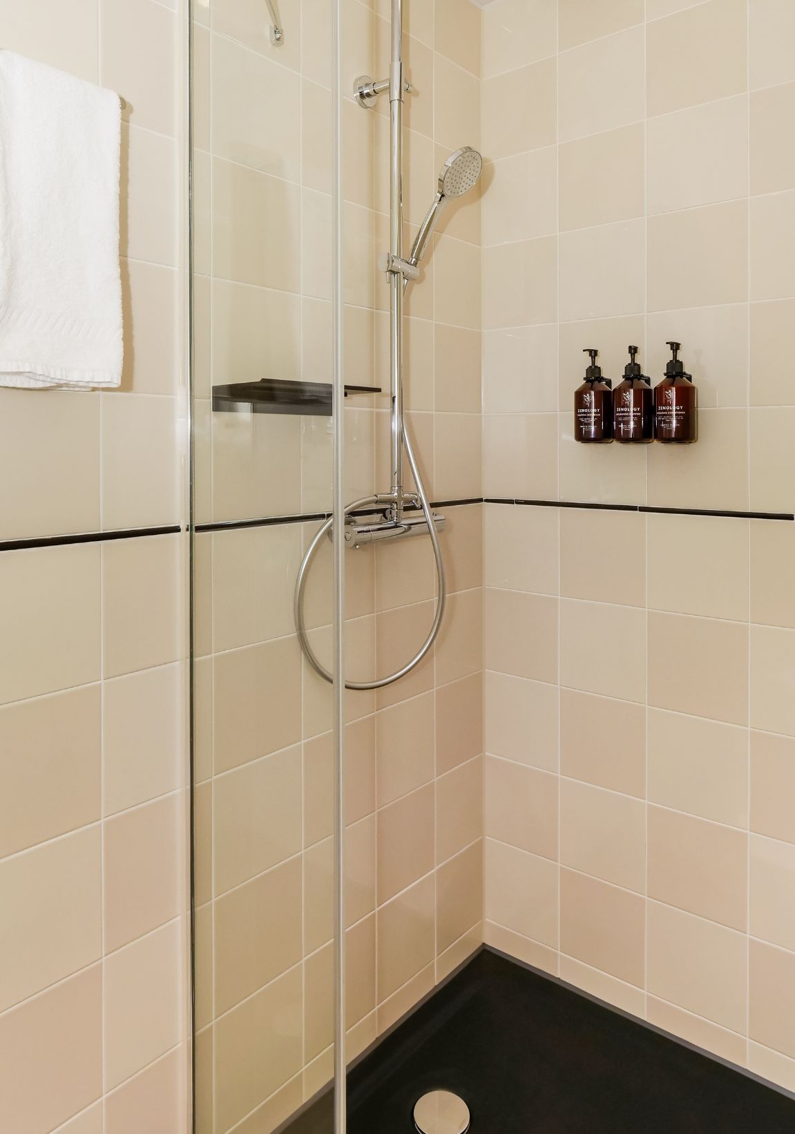 Un baño con mampara de vidrio, ducha de mano y grifo empotrado. Hay tres botellas de champú en la pared.