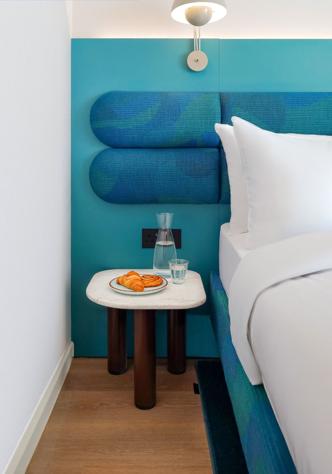 A signature room with a teal accent wall, a white bed, a small table with a croissant and glasses, and a lamp.
