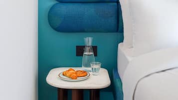 A signature room with a teal accent wall, a white bed, a small table with a croissant and glasses, and a lamp.