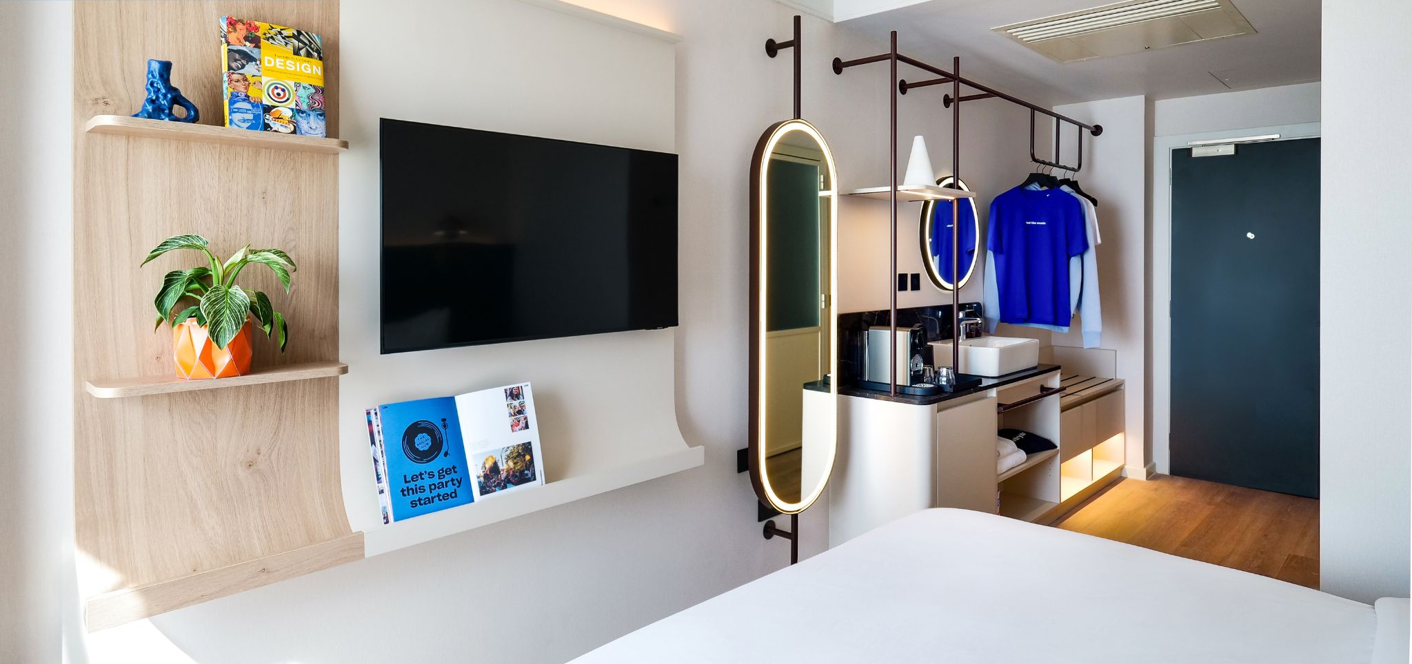 Una habitación de hotel moderna con una televisión de pantalla plana, un espejo grande, un tocador con lavabo y una cama. Hay repisas con objetos decorativos y un letrero en la pared.