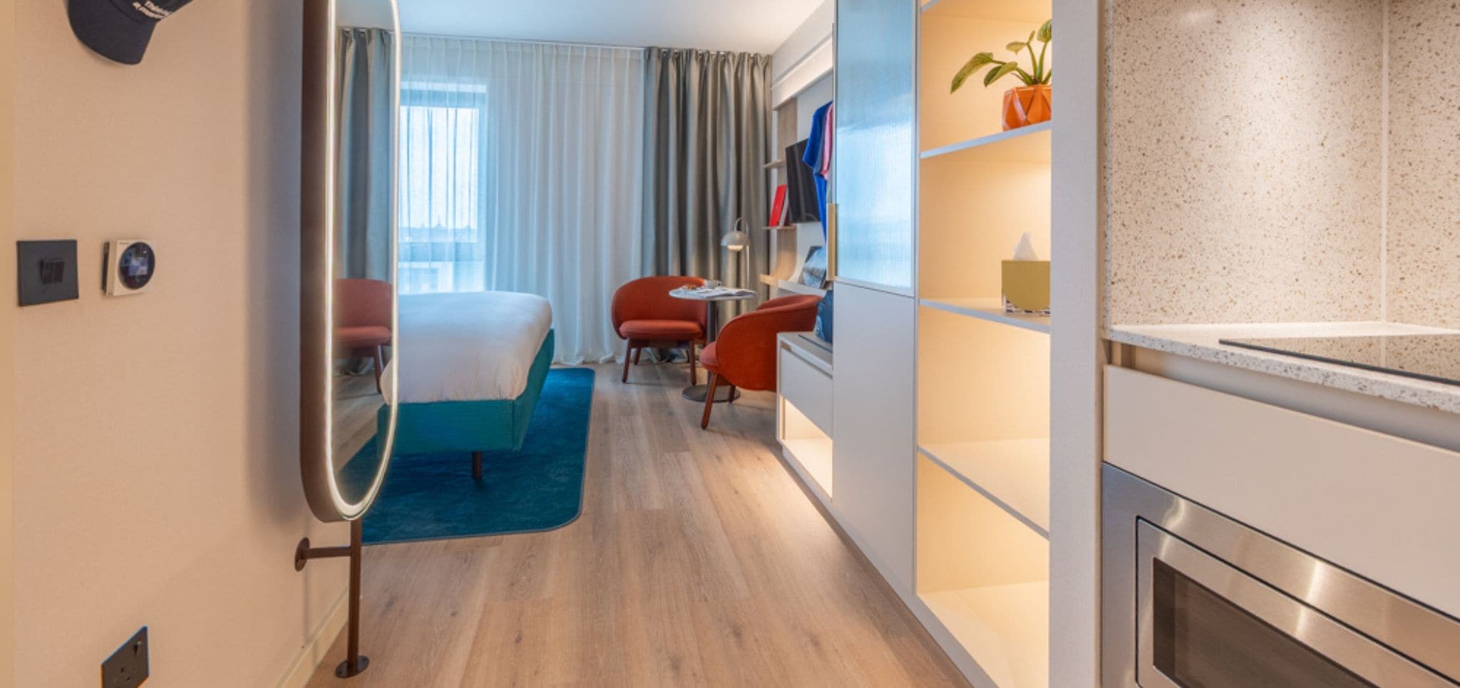 A modern hotel room with a bed, two red armchairs, a small table, a mirror, a potted plant, and a kitchenette.