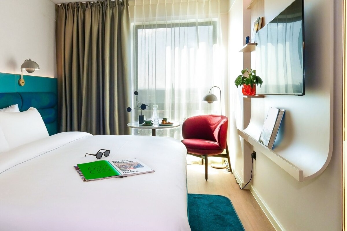 A hotel room with a bed, a desk, a chair, a TV, and a window with curtains.
