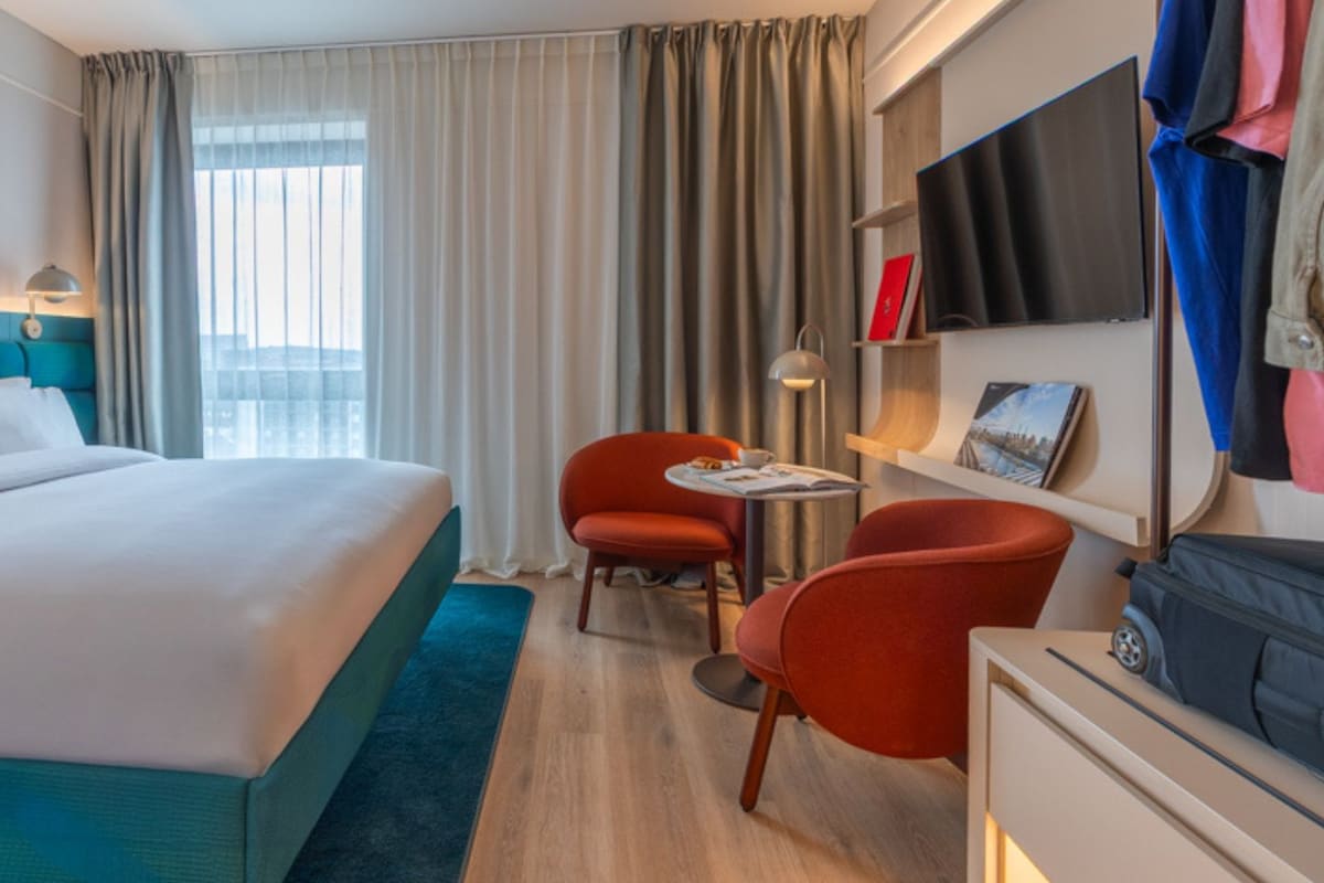 A hotel room with a large bed, two red chairs, a small table, a TV, and a wardrobe.