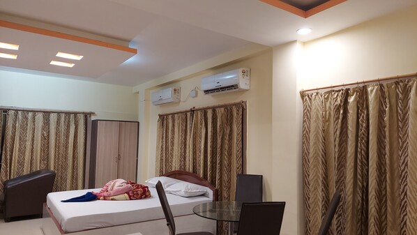Une chambre d’hôtel avec un lit, une chaise, une table et une armoire. Il y a des rideaux aux fenêtres et un appareil de climatisation fixé au mur.