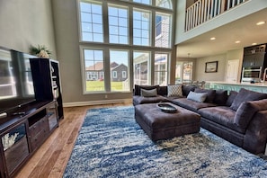 A spacious living room with a large window, a dark brown leather sofa, a TV on a wooden stand, and a kitchen in the background.