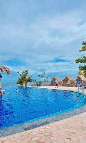 A swimming pool with clear blue water, surrounded by thatched-roof huts and palm trees.