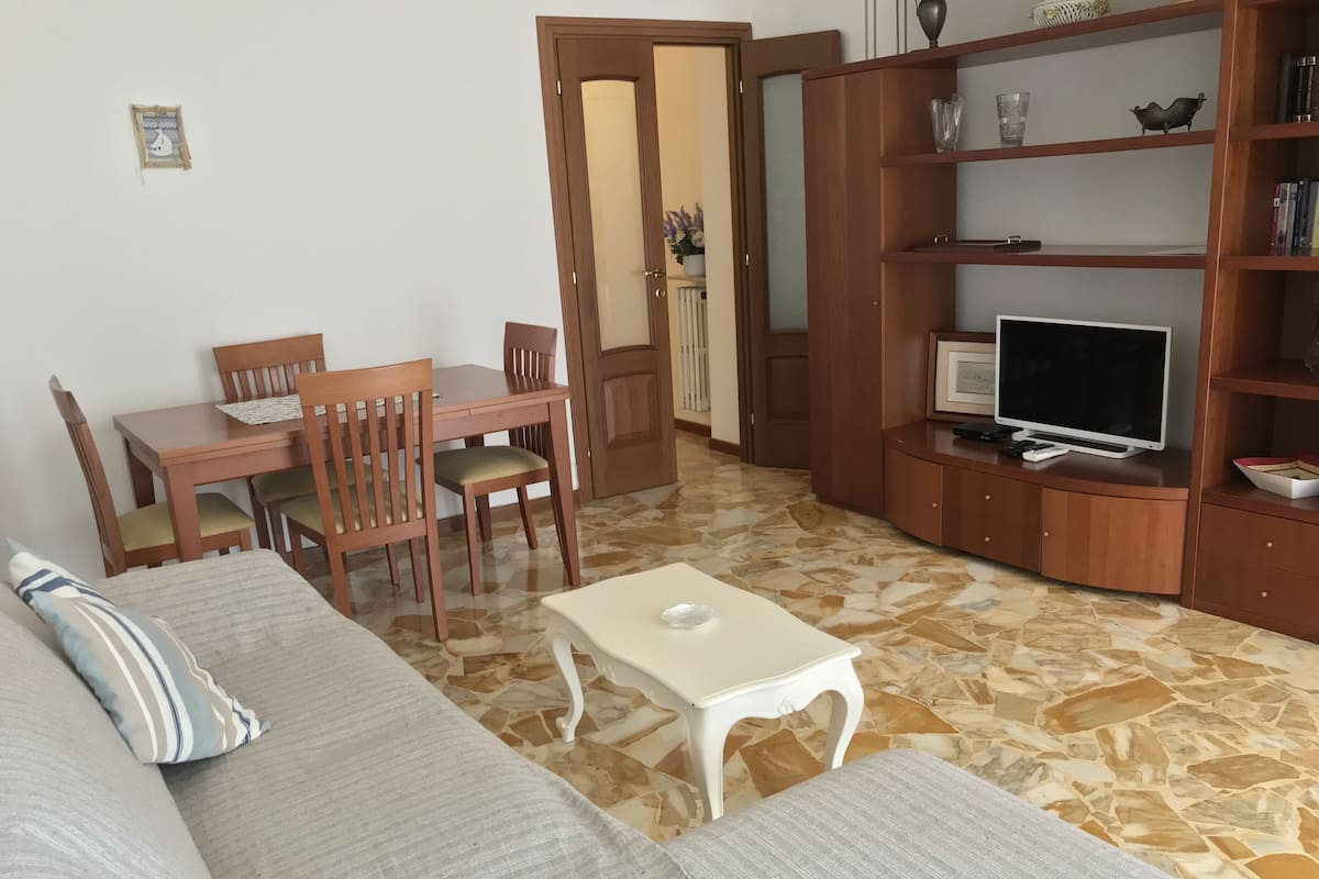 Ein Wohnzimmer mit einer Couch, einem Esstisch mit Stühlen, einem Fernseher auf einer Holzkommode und einem gemusterten Boden.
