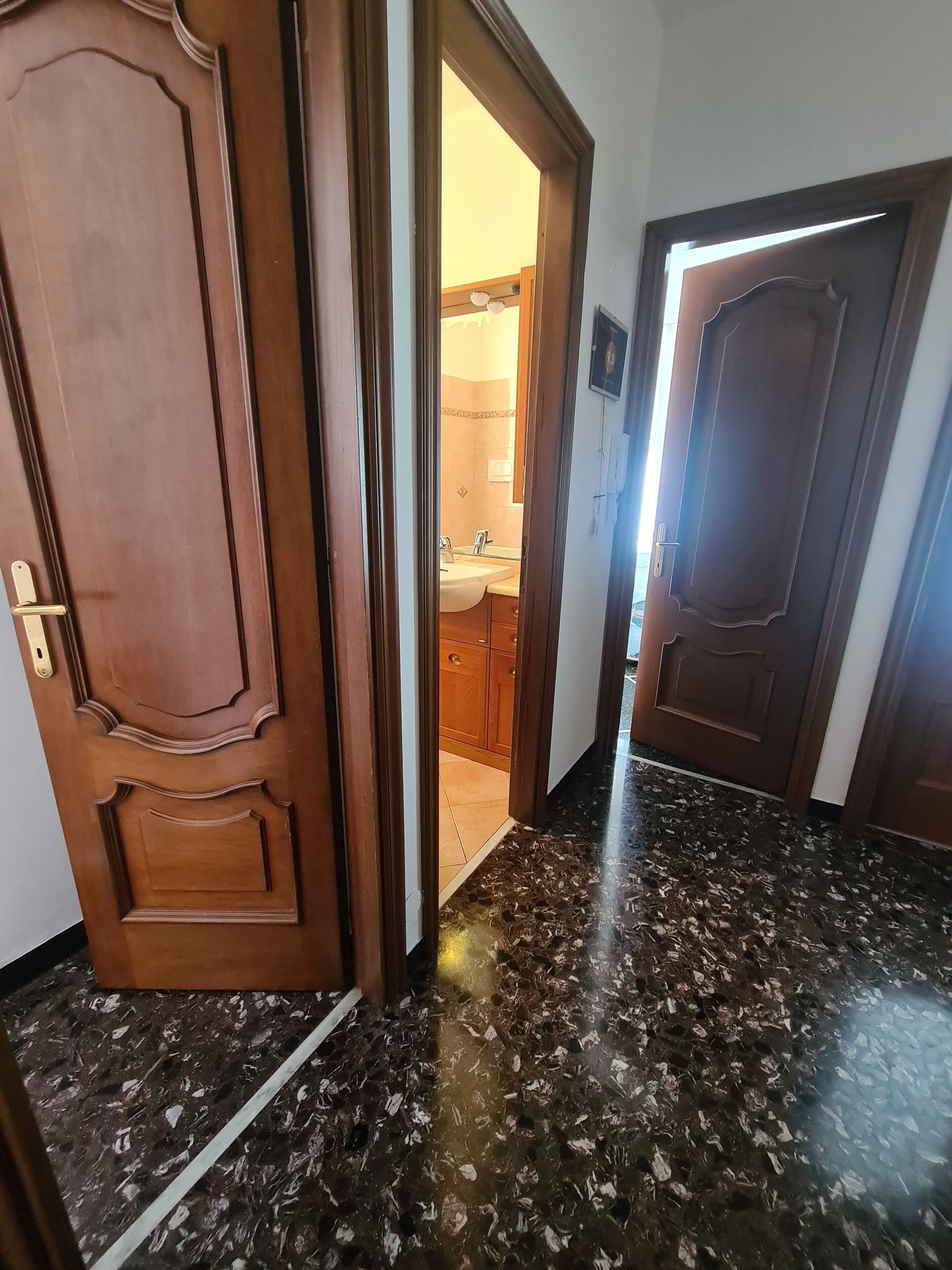 Un couloir avec une porte en bois, une salle de bain visible à travers une porte ouverte et un sol en marbre foncé.