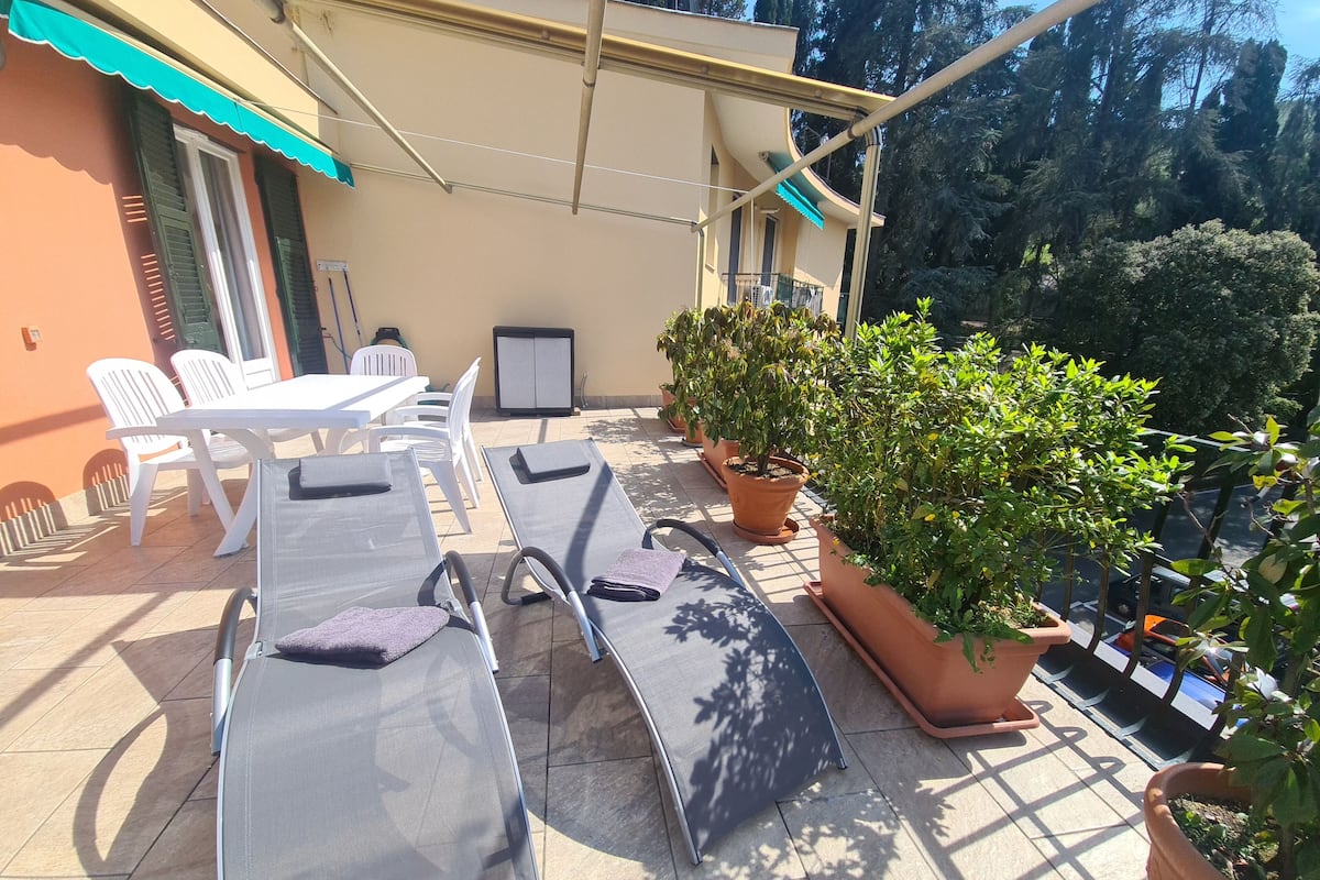 Ein sonniger Außenbereich mit Sitzgelegenheiten, Lounge-Stühlen und einem Tisch, umgeben von Blumentöpfen und einem Gebäude mit blauem Markisen.