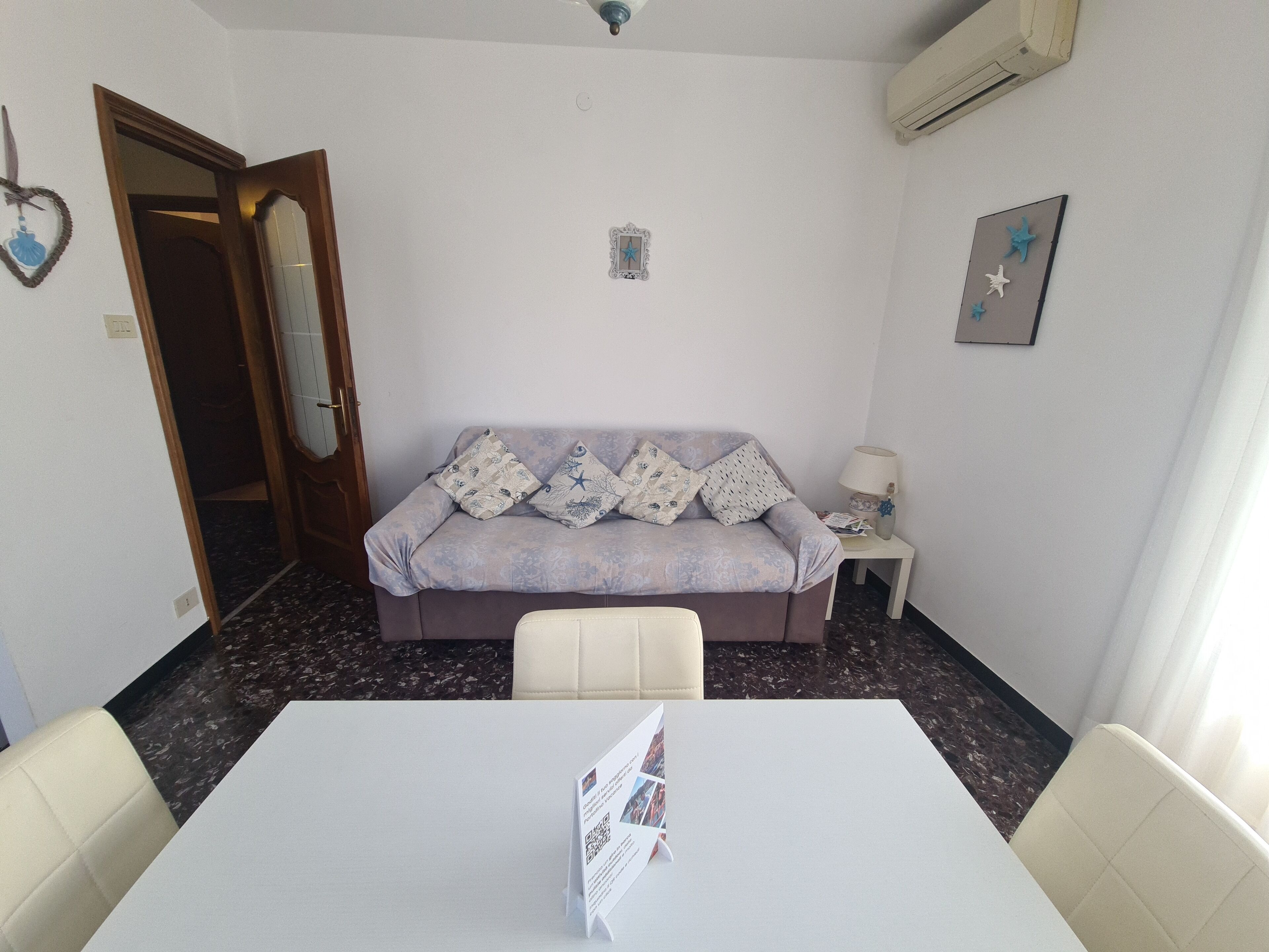 Una habitación pequeña y bien iluminada con sofá, una mesa y una silla. Se ve una puerta que da a otra habitación, una mesita con lámpara y un aire acondicionado instalado en la pared.