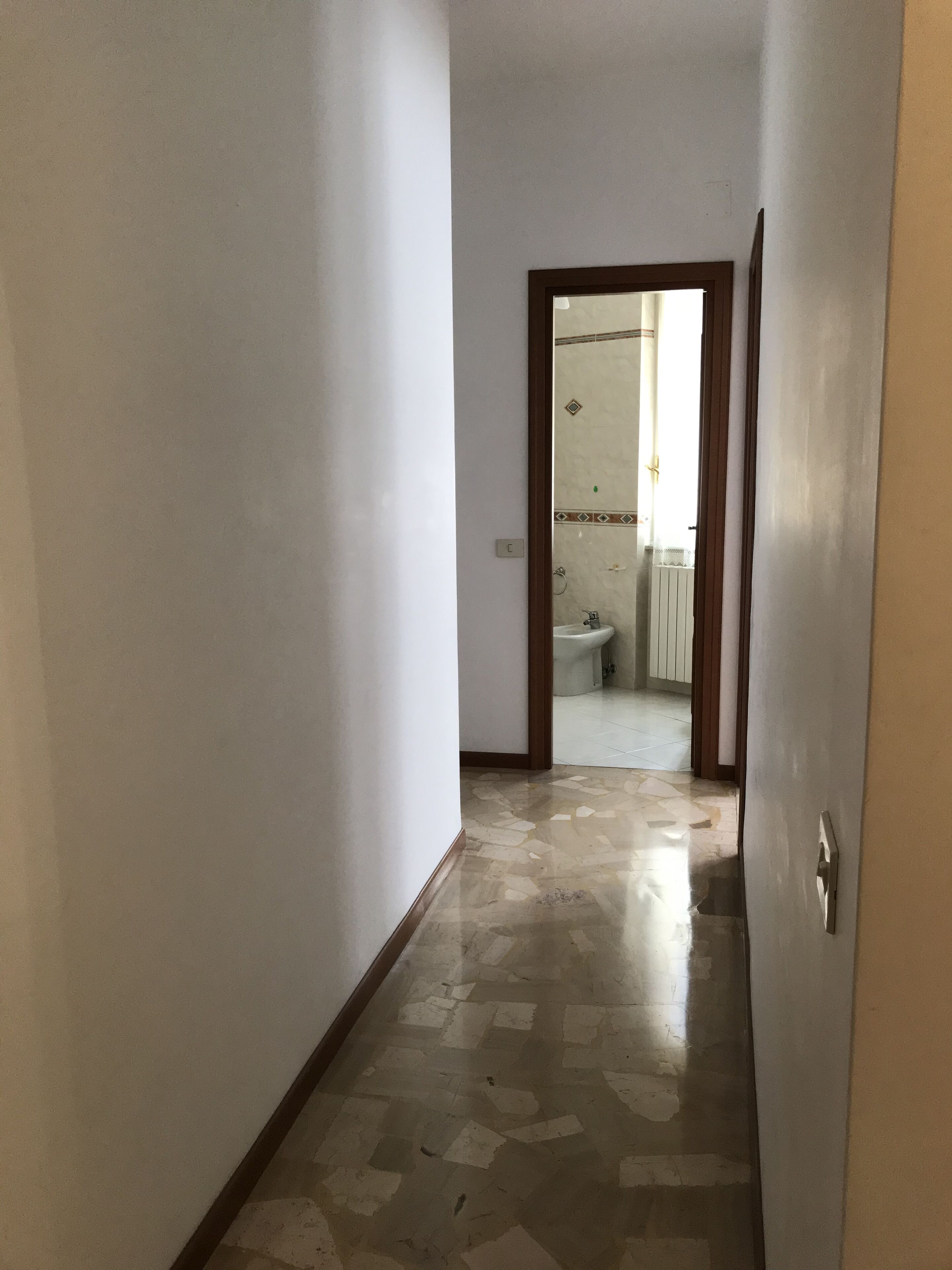 Ein schmaler Korridor mit Fliesenboden, der zu einem Badezimmer mit Toilette und Waschbecken führt.