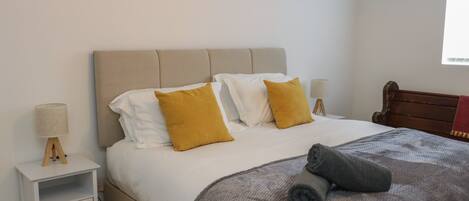 A neatly made bed with a grey blanket and two pillows, a bedside table with a lamp, and a wooden chair.