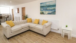 A living room with a beige sectional sofa, wooden flooring, a painting of a sailboat on the wall, and a small white table with a plant. - The Church (Holyhead)