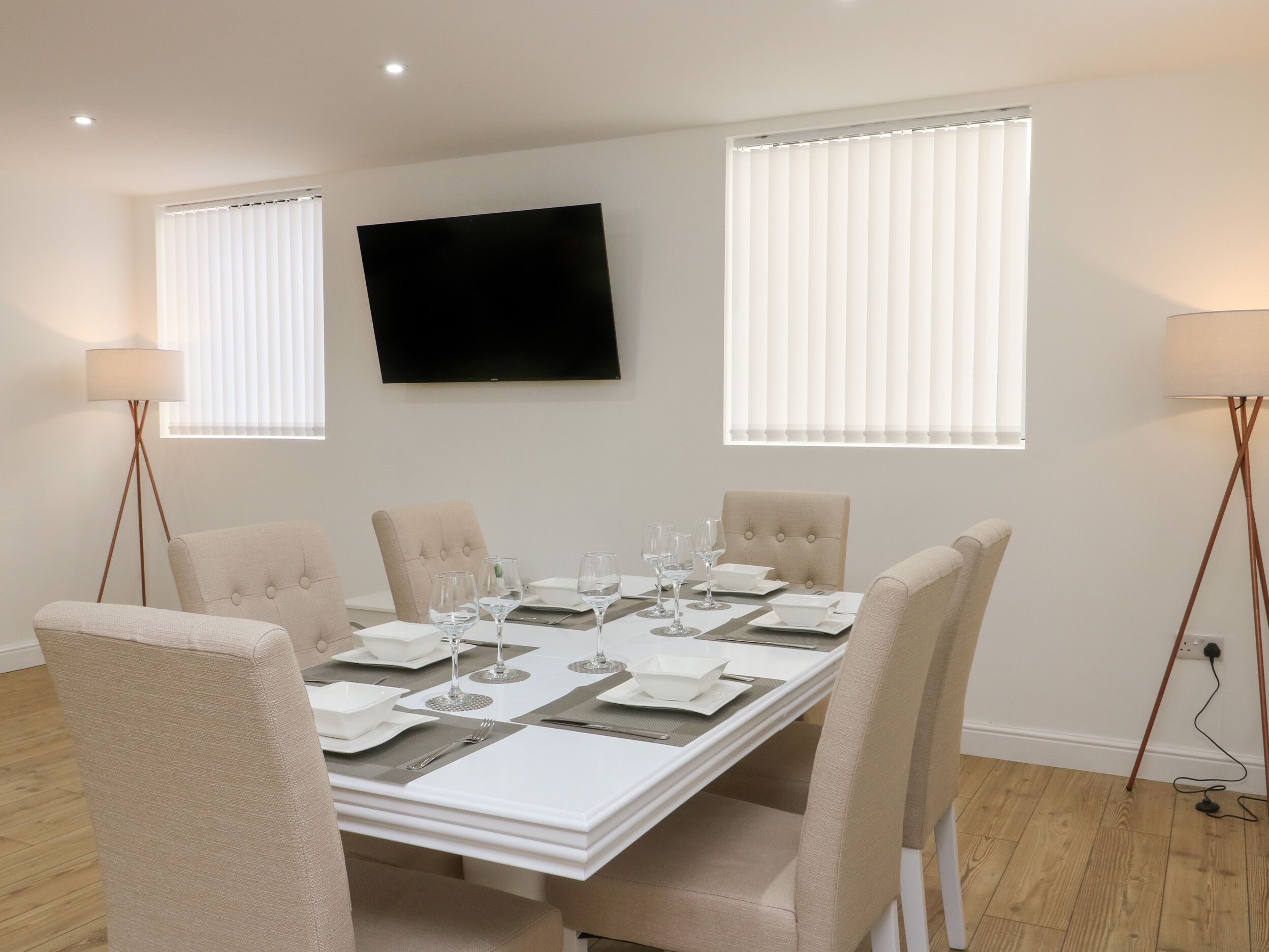A dining room with a rectangular table set for six, flanked by two floor lamps, and a flat-screen TV mounted on the wall.