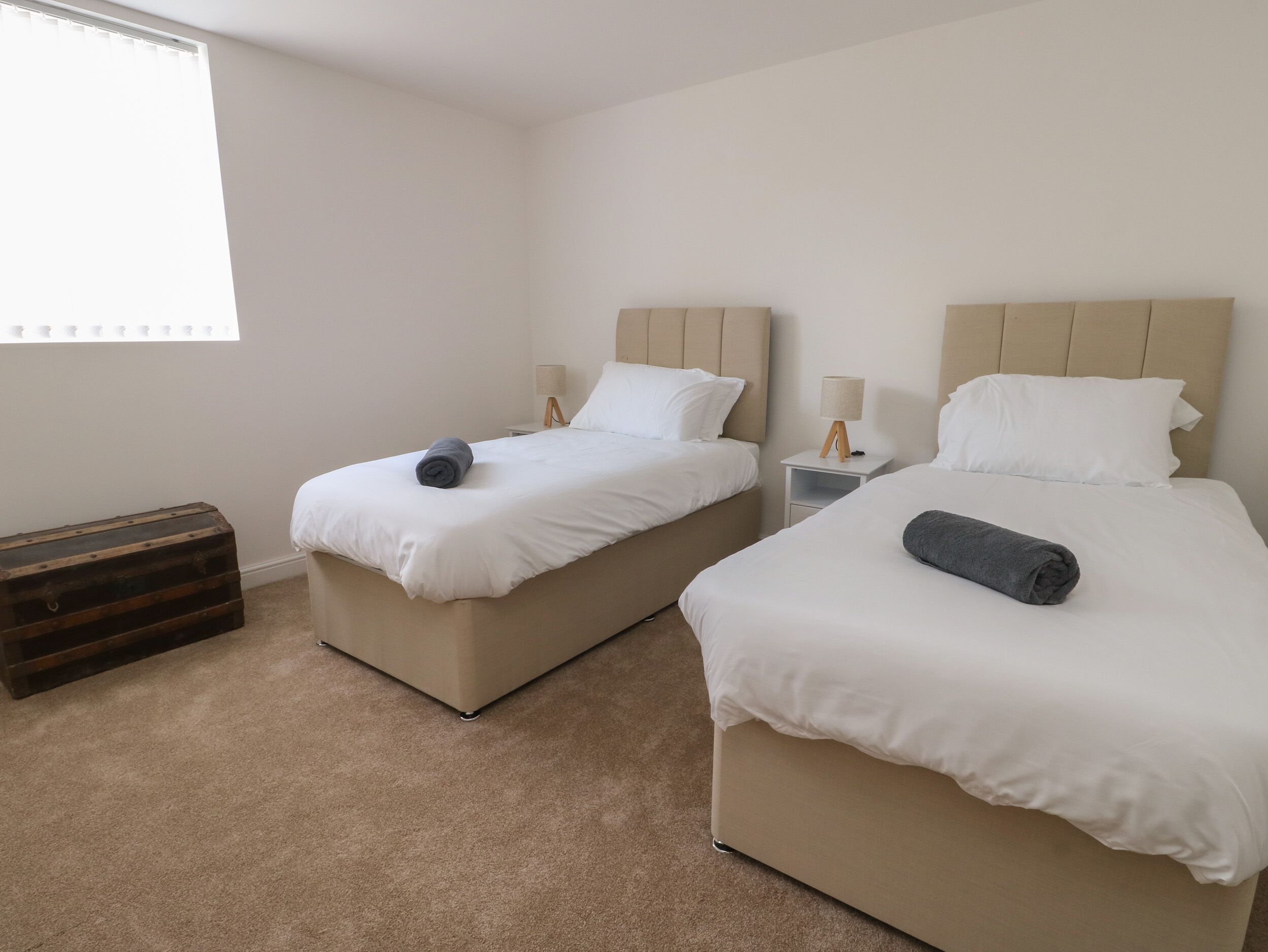 A hotel room with two beds, each with a headboard, a bedside table with a lamp, and a window with blinds.