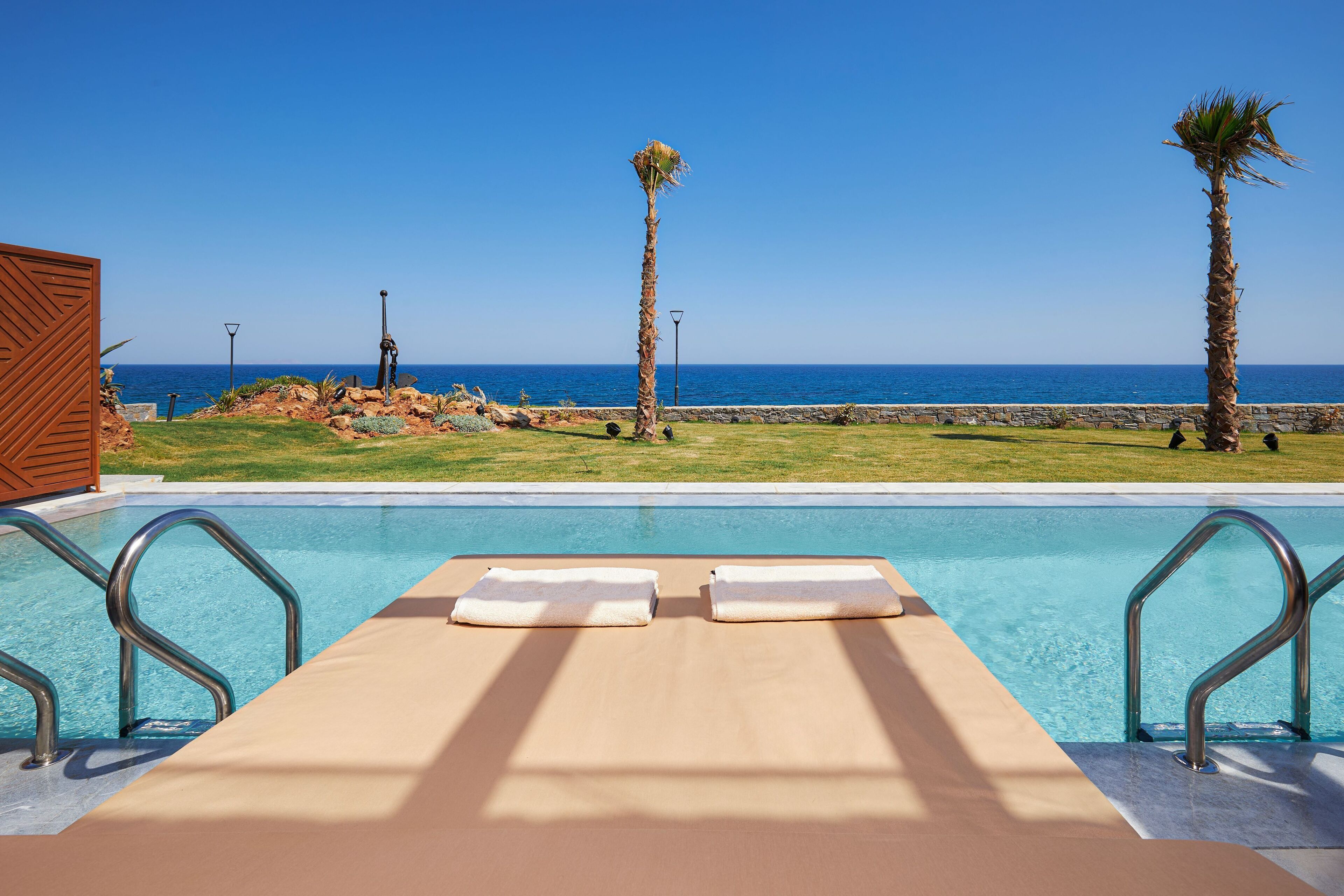 Un espace aménagé au bord de la piscine avec deux chaises longues, une vue dégagée sur l’océan et un mur de pierre en arrière-plan.