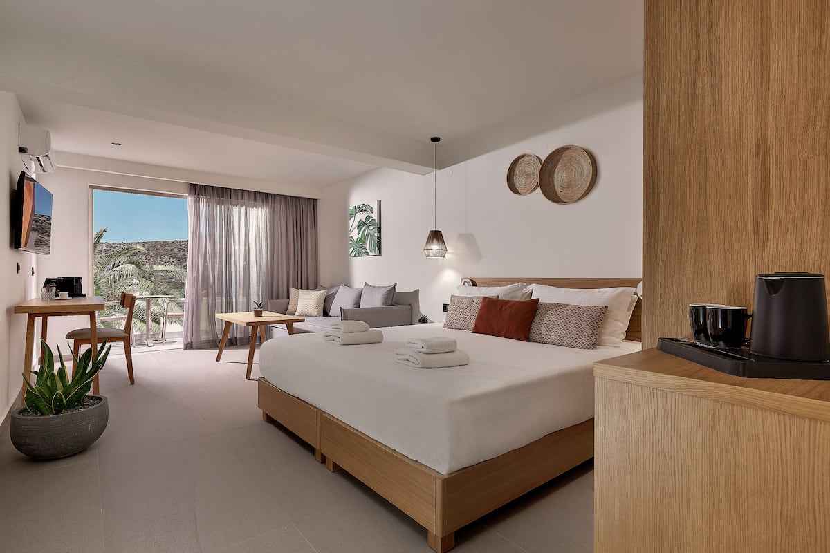 Een moderne hotelkamer met een groot bed, een klein bureau, een bank en een balkon met uitzicht op palmbomen.