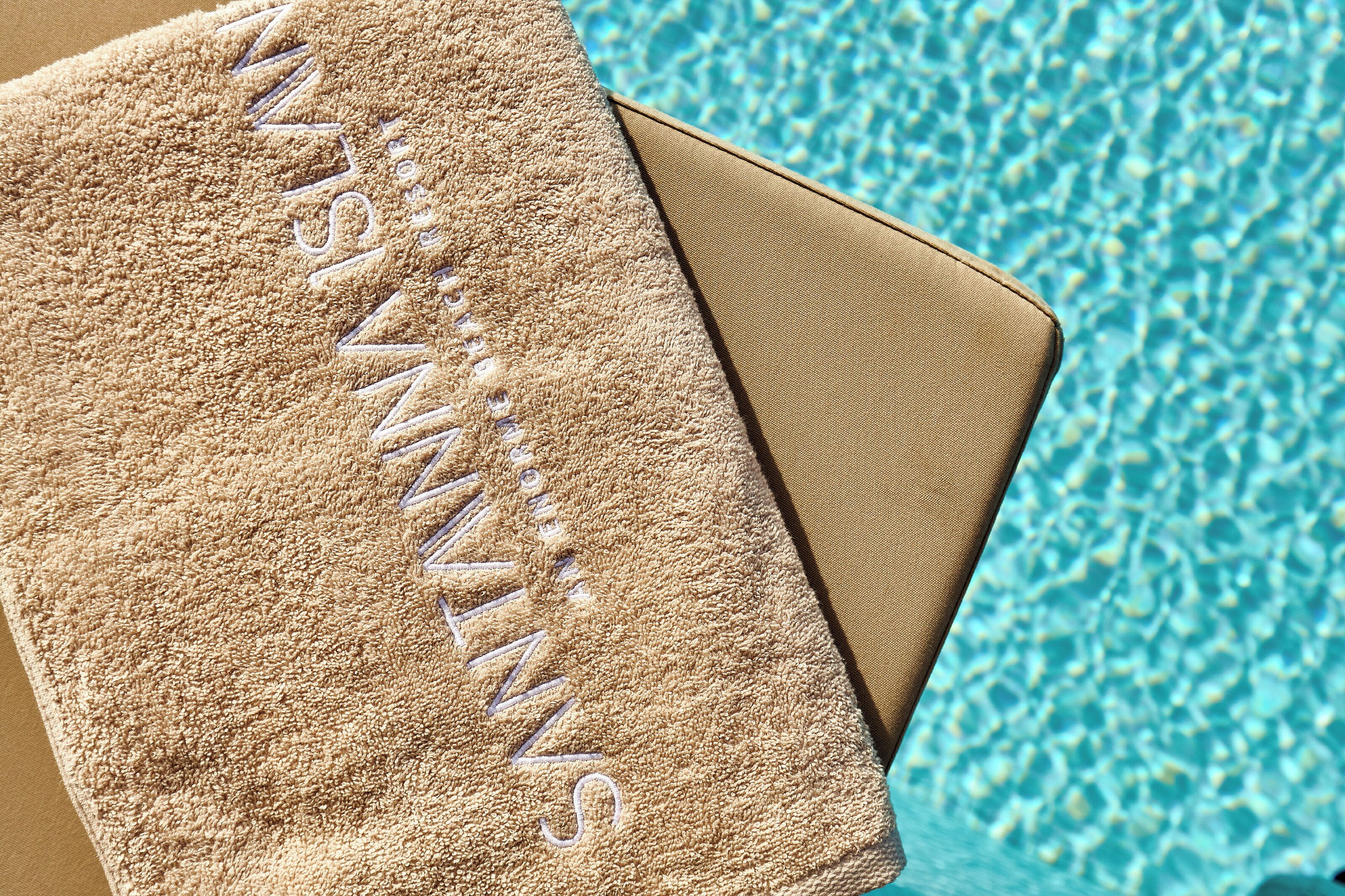 A towel with the word "MANN'S" embroidered on it, placed next to a pool with clear blue water.