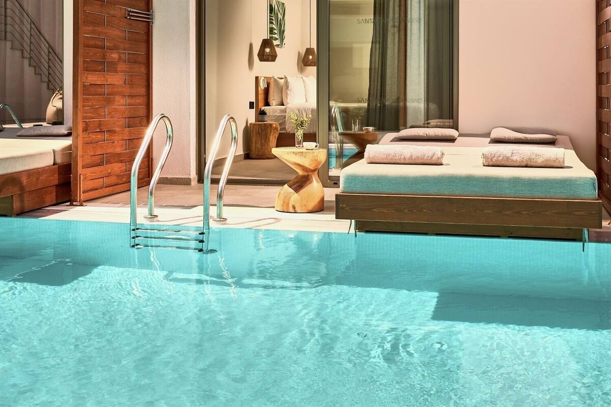 Een zwembad met een houten terras, een zithoek met een bank en kussens, en een glazen deur die naar een kamer leidt.