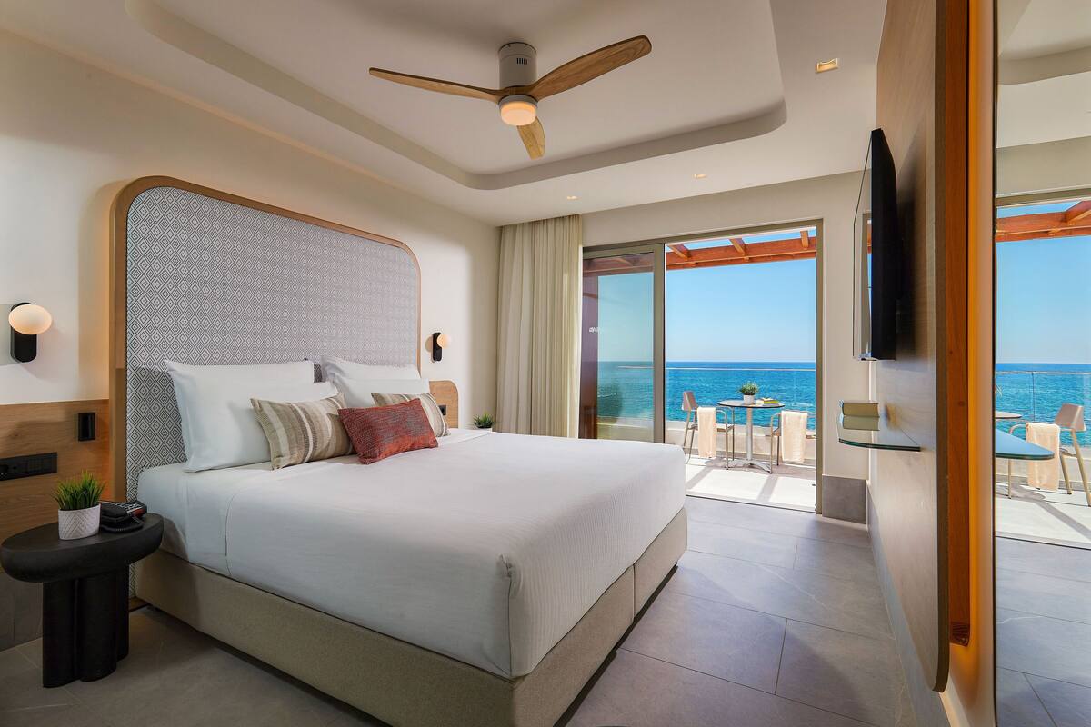 Een moderne hotelkamer met een groot bed, een plafondventilator, een televisie en een balkon met uitzicht op zee.
