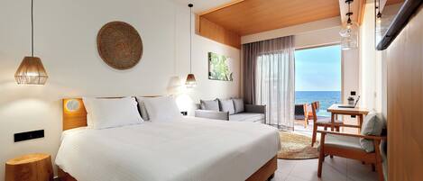 A modern hotel room with a large bed, a wooden headboard, a round wall decoration, a desk with a laptop, and a view of the sea through large windows.