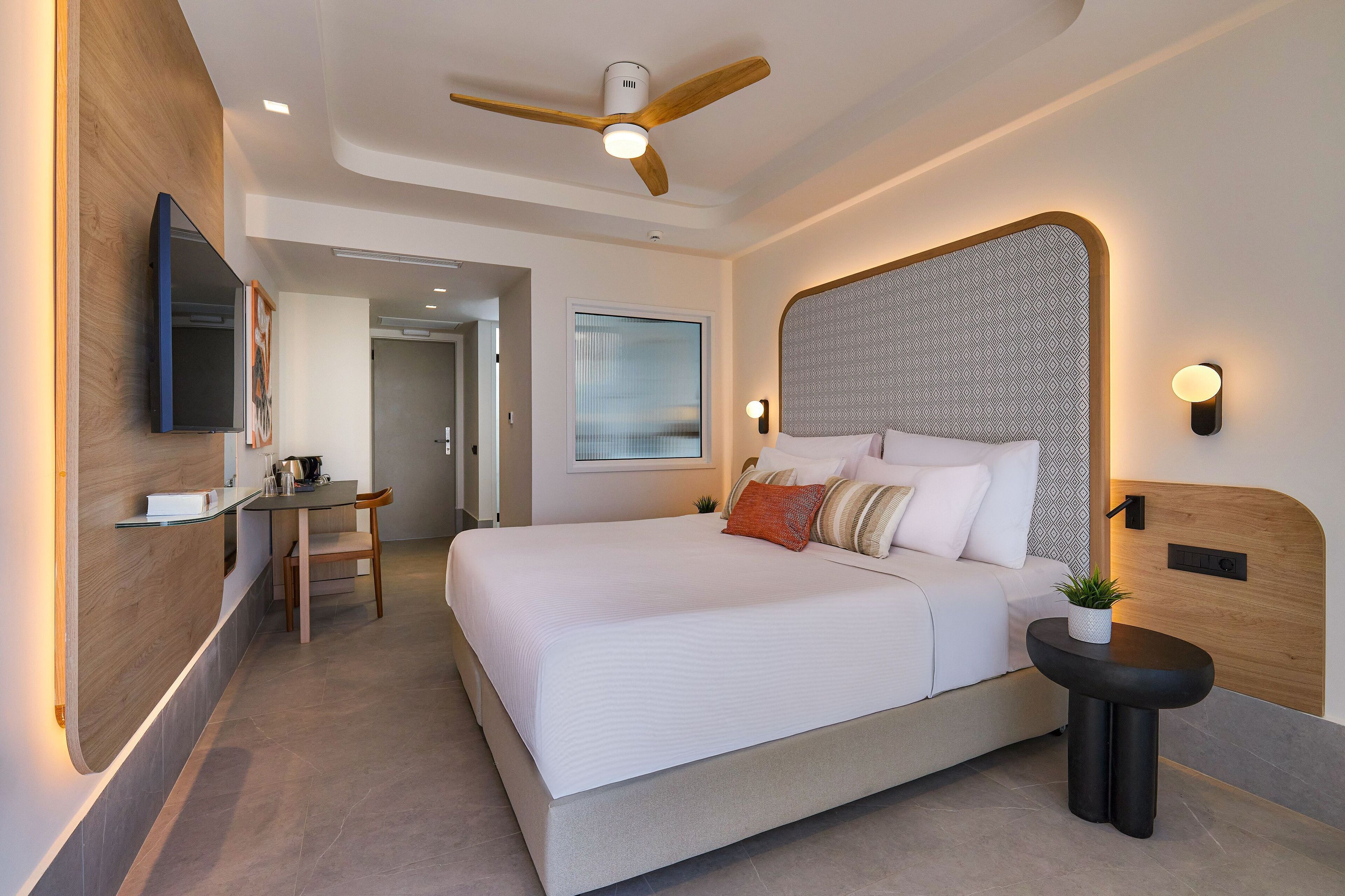 A modern hotel room with a large bed, a ceiling fan, a small table with a plant, and a desk with a chair.