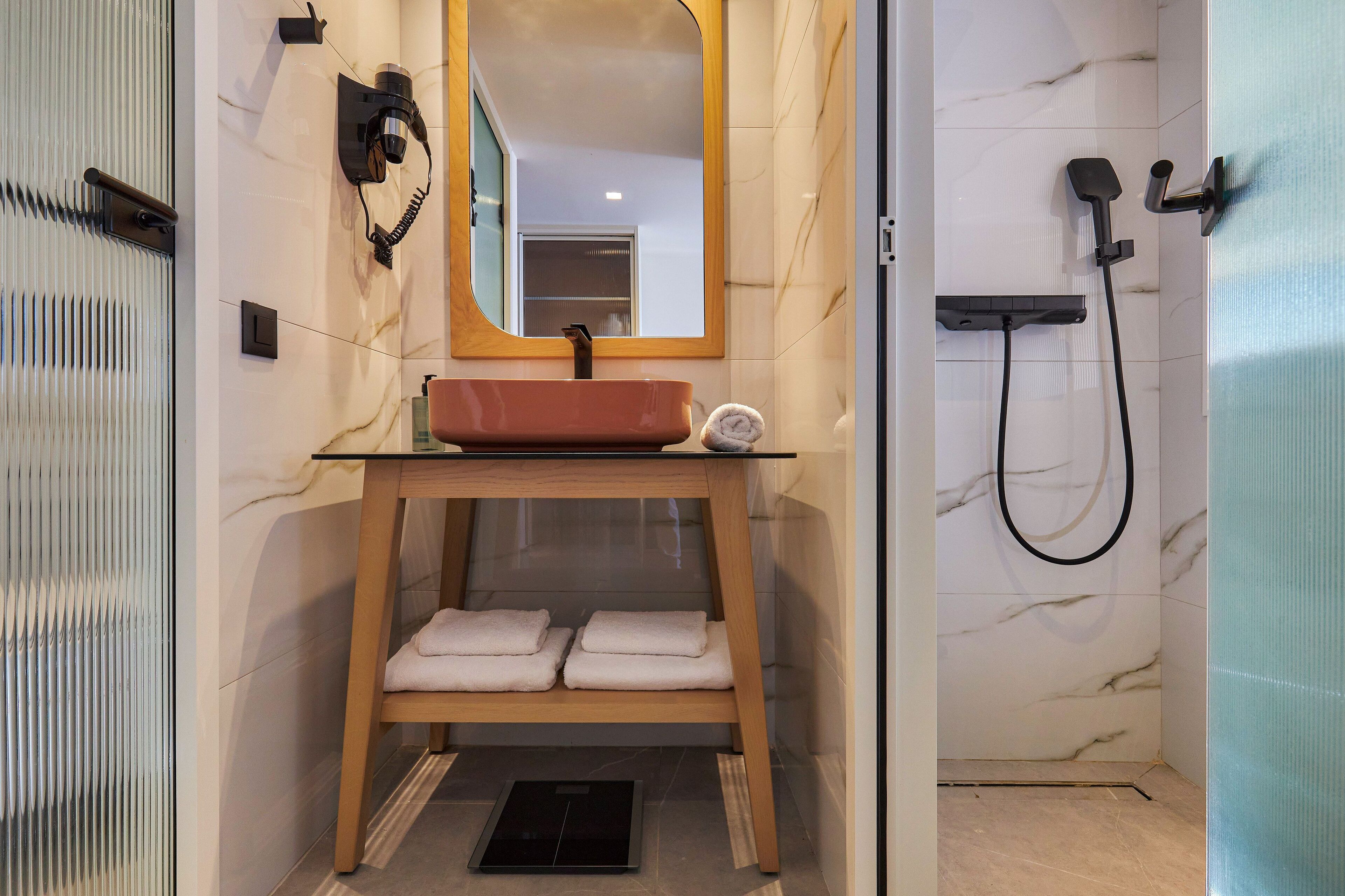 Un baño con pared de mármol, tocador de madera, lavamanos, espejo, perchero para toallas y zona de ducha.