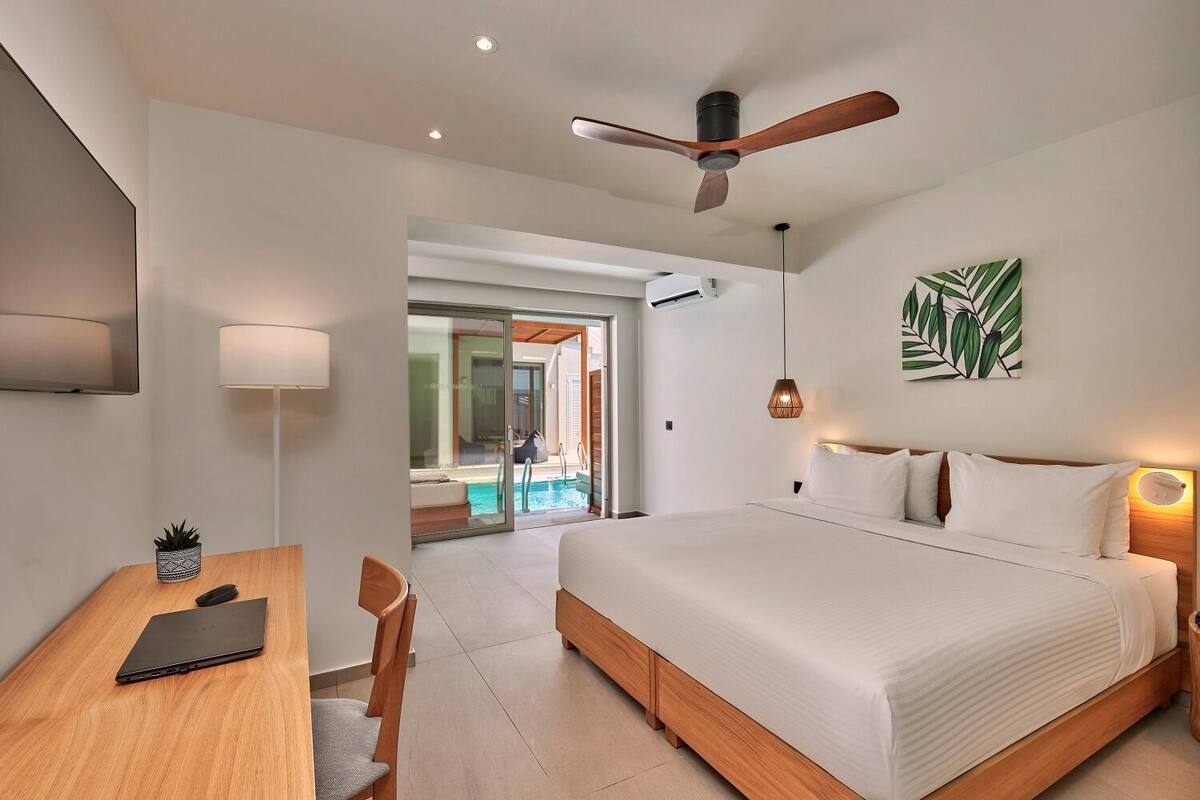 Een moderne hotelkamer met een groot bed, een bureau met een laptop, een plafondventilator en uitzicht op een zwembad.