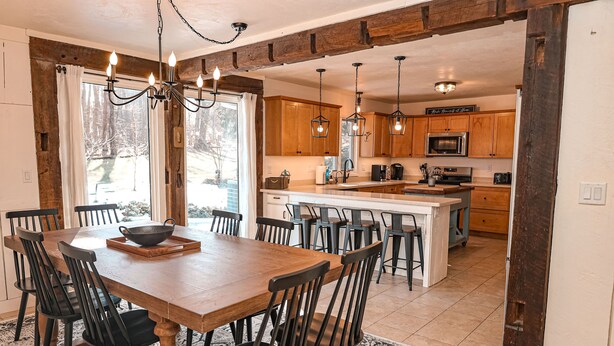 A dining area with a wooden table and chairs, a chandelier, and a view of the kitchen in the background. - Family Friendly Retreat, sleeps 12+, full basketball court, Large inground pool (La Porte)