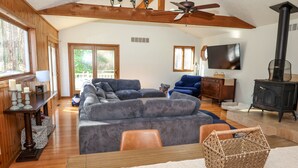 A cozy living room with a large gray sectional sofa, a wooden ceiling fan, a black wood-burning stove, and a television mounted on the wall. - Family Friendly Retreat, sleeps 12+, full basketball court, Large inground pool (La Porte)