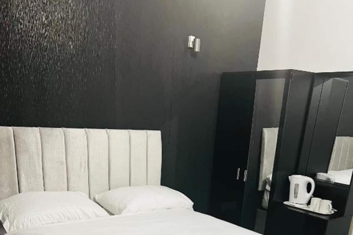 Ein ordentlich bezogenes Bett mit weißen Leinen und einem gefalteten Handtuch, ein dunkler Holzschrank und ein kleiner Tisch mit Wasserkocher und Tassen.