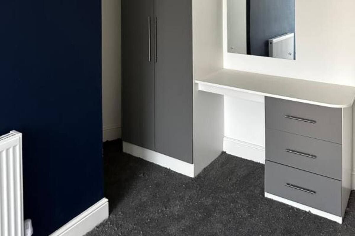 A bedroom with a dark blue wall, a grey wardrobe, a white dresser, and a ceiling light.