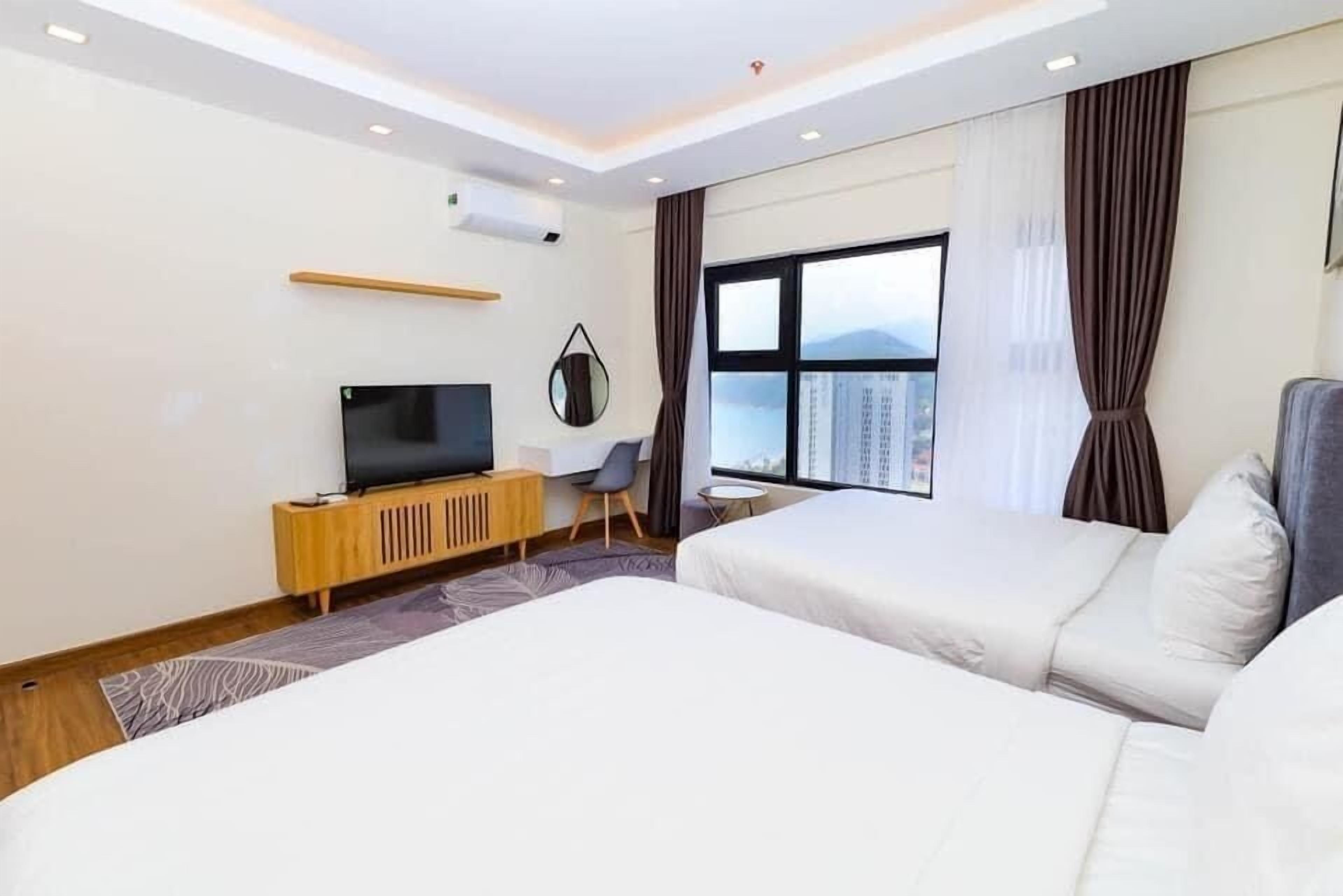 A hotel room with two beds, a TV on a wooden cabinet, a small table, and a view of the city through large windows.