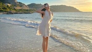 Una dona amb un vestit blanc dempeus a la platja, amb ones, un cel clar i un edifici al fons.