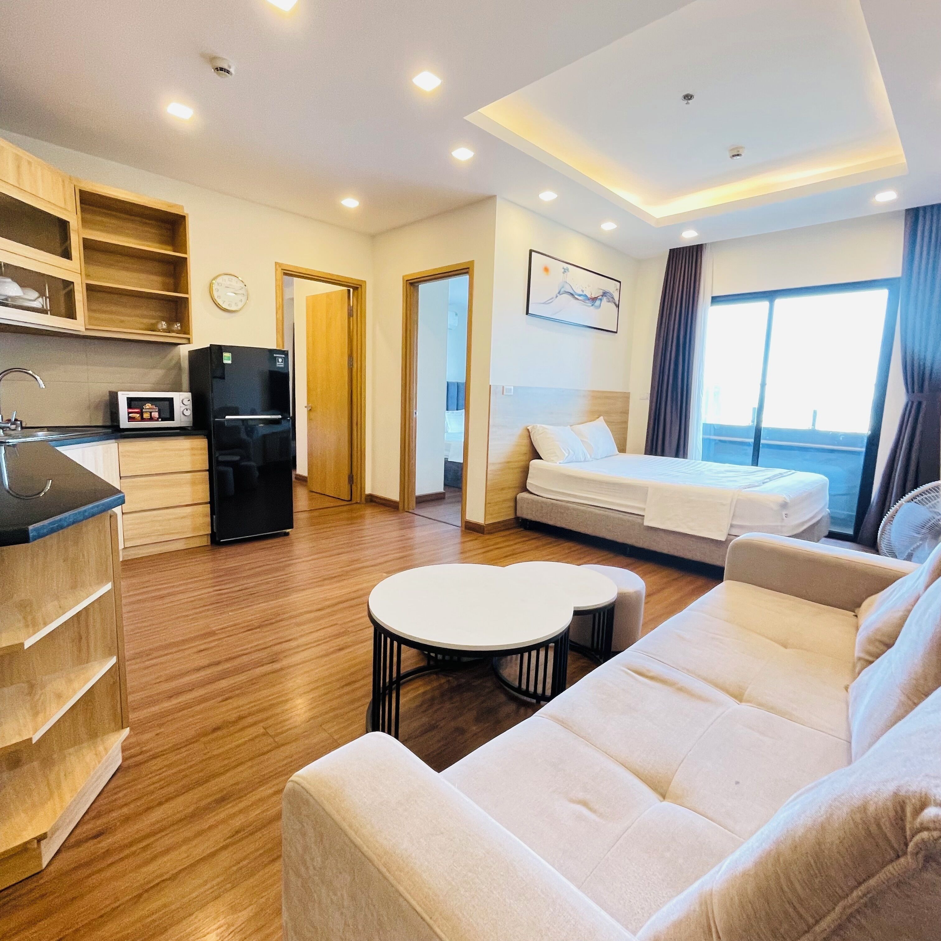 A modern hotel room with a kitchenette, a sofa, a bed, and a large window.