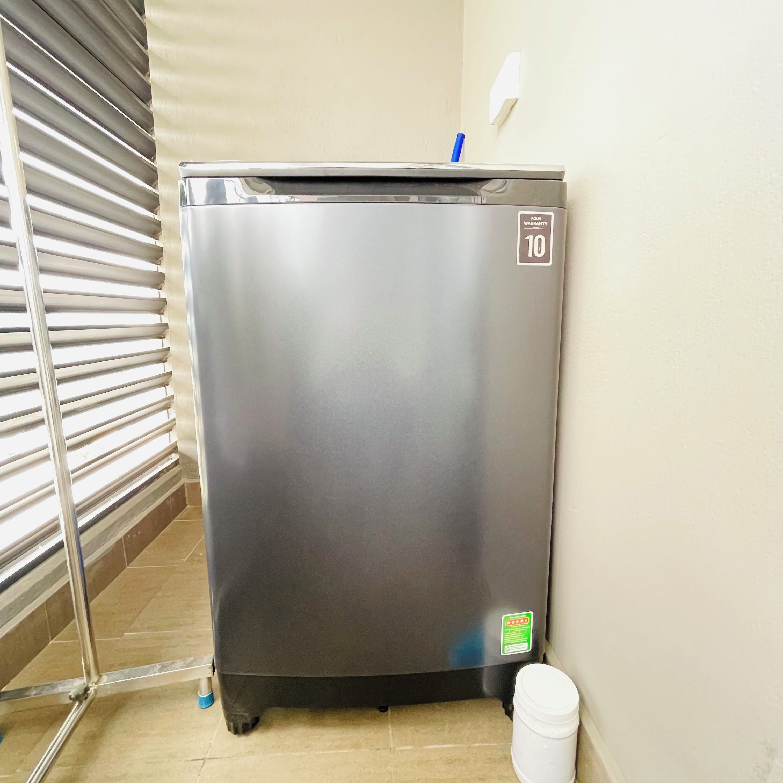 Refrigerador de acero inoxidable compacto con una etiqueta que indica una garantía de 10 años, ubicado en un rincón de una habitación con piso de baldosas y un estante metálico.