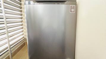 Refrigerador de acero inoxidable compacto con una etiqueta que indica una garantía de 10 años, ubicado en un rincón de una habitación con piso de baldosas y un estante metálico.