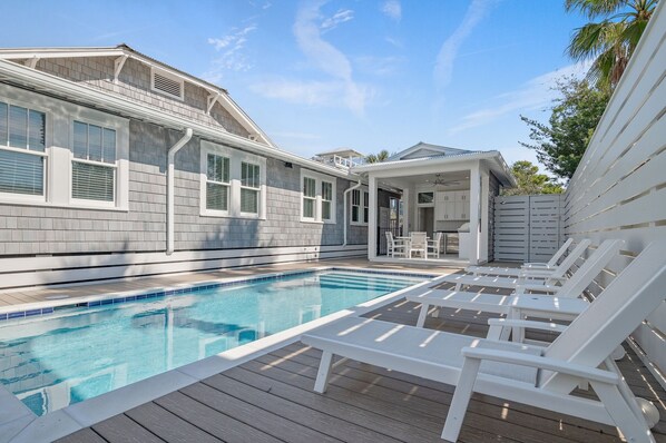 A swimming pool with white lounge chairs, a covered patio, and a house with grey siding and white trim.