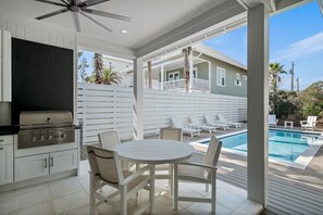 An outdoor kitchen with a grill, a pool, and a seating area with white chairs and a table.