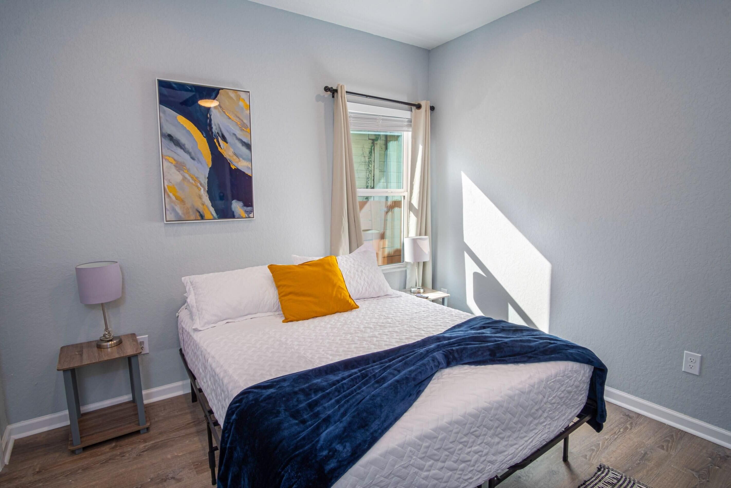 A bedroom with a bed, a nightstand, a lamp, a window with curtains, and a painting on the wall.