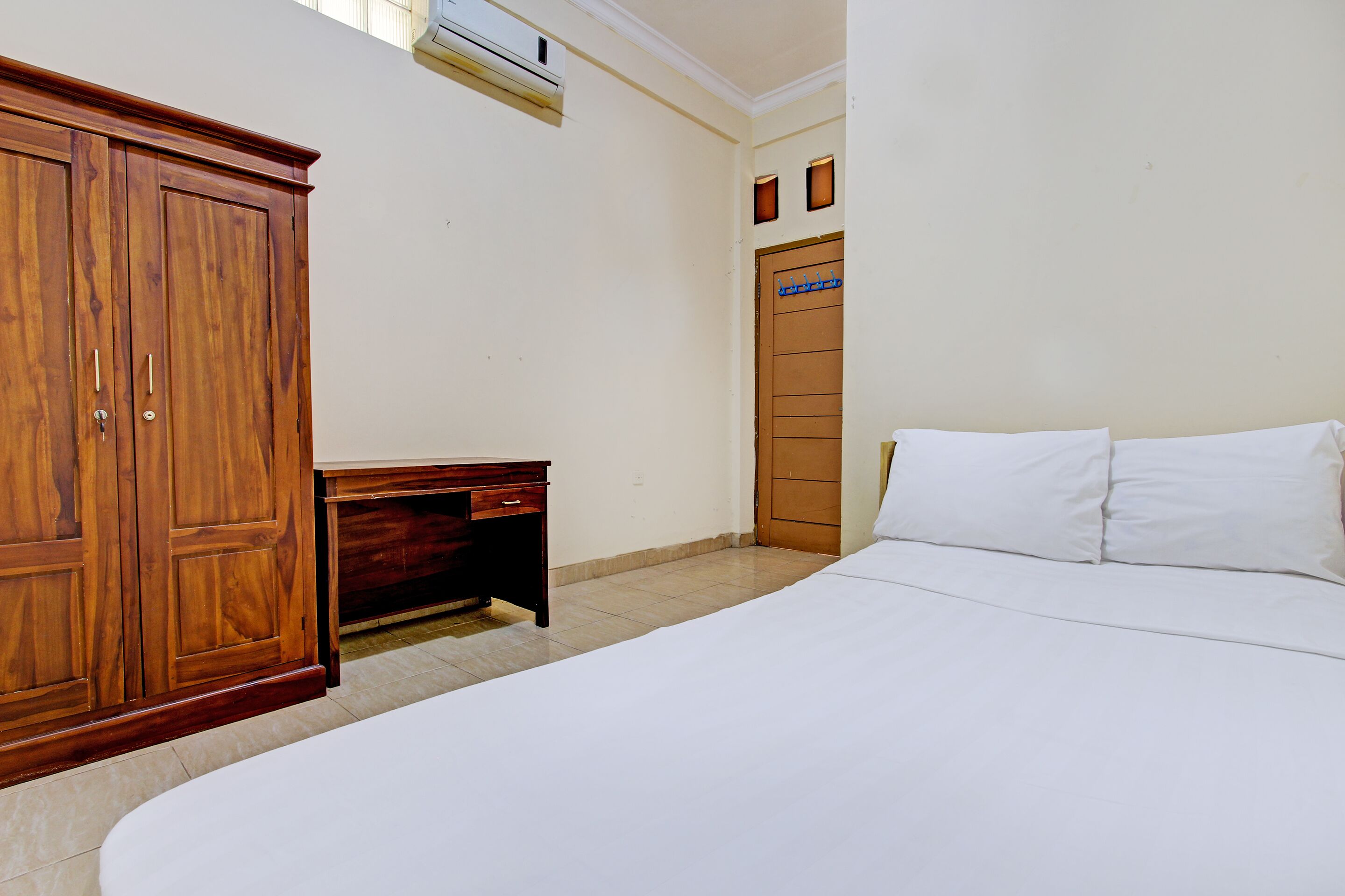 A bedroom with a large wooden wardrobe, a small desk, a bed with white linens, and a door with a blue sign.