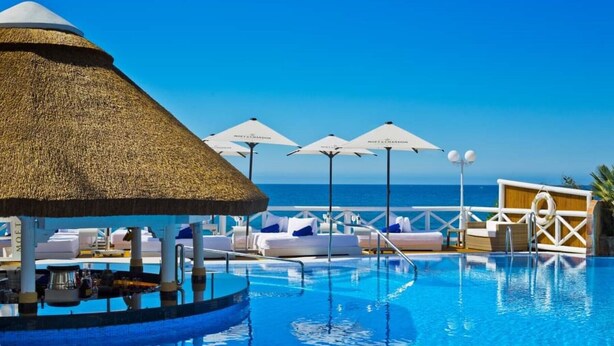 A pool area with lounge chairs and umbrellas, a thatched roof structure, and a view of the sea.