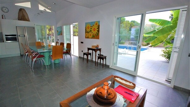 A spacious living area with a dining table, chairs, a kitchen area, and a large sliding glass door leading to a pool. - SAONA VILLAS (VN OLIVE): villa for 10 people, private swimming pool, air co (Les Tres Cales)