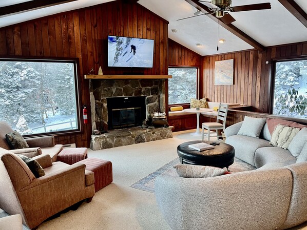A cozy living room with a stone fireplace, wooden walls, and large windows offering a view of snow-covered trees. - SnowMonte in the Pines

 (Dunmore)