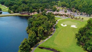 An aerial view of a golf course with a lake, surrounded by dense forest, and a parking area with buildings.