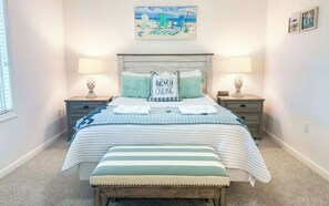 A bedroom with a bed, two bedside tables, a headboard, a bench, and a beach-themed wall art.