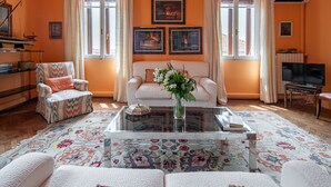 Un salon comprenant un canapé, un fauteuil et une table basse. Il y a deux fenêtres avec des rideaux, un tableau encadré au mur et un téléviseur posé sur un meuble.